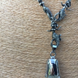 May include: A silver necklace with a bell pendant and multiple mermaid charms. The necklace is on a brown textured surface.