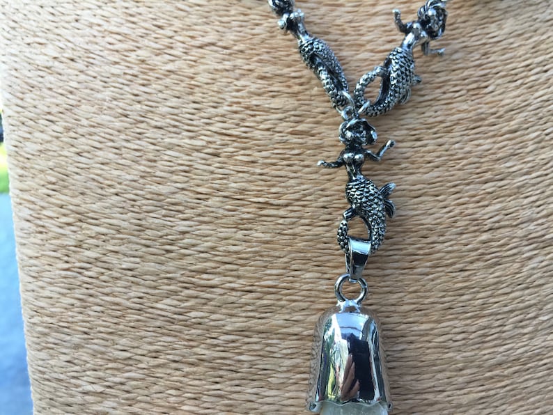 May include: A silver necklace with a bell pendant and multiple mermaid charms. The necklace is on a brown textured surface.