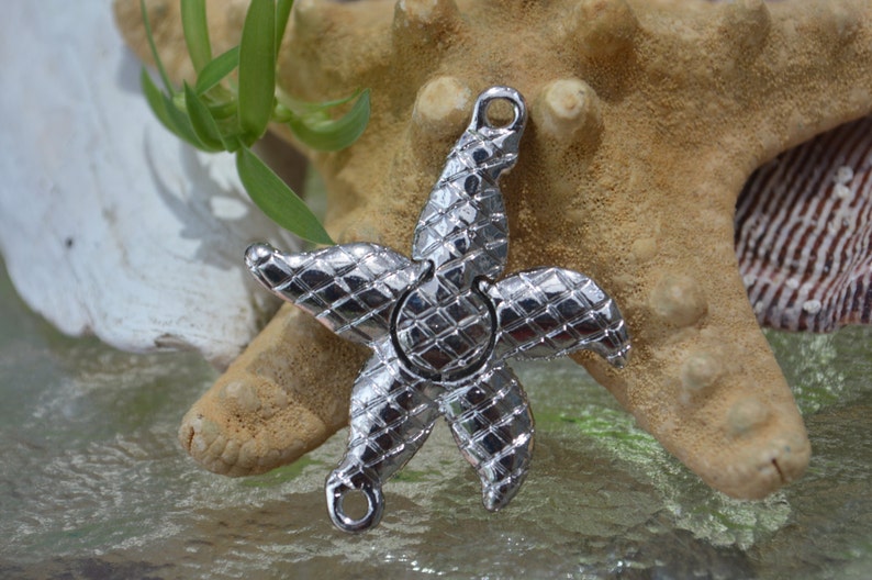 May include: A silver-toned metal starfish charm with a textured surface. The charm has two loops for attaching to jewelry or other crafts.