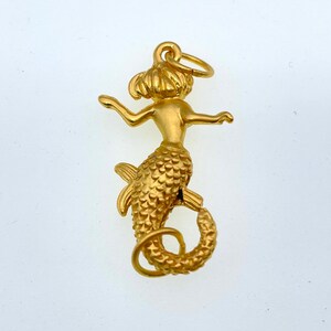 May include: A gold-colored mermaid charm with a detailed, textured tail and a loop for attaching to a necklace or bracelet.