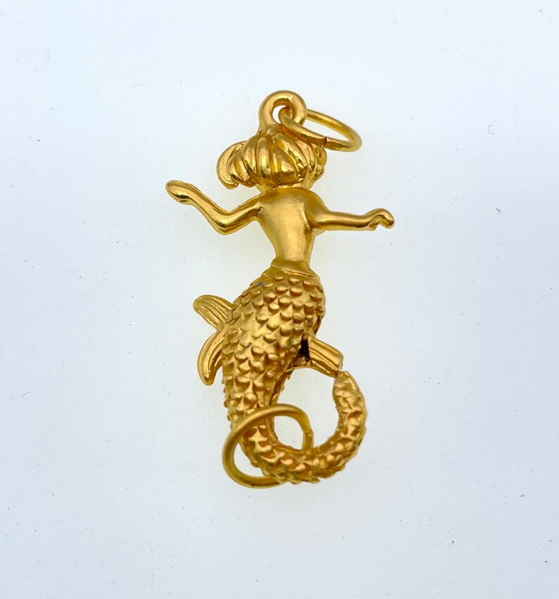 May include: A gold-colored mermaid charm with a detailed, textured tail and a loop for attaching to a necklace or bracelet.