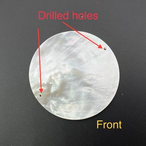 May include: A round, iridescent white shell with two drilled holes and the word "Front" in yellow. The shell has a pearlescent sheen and a smooth surface. The drilled holes are small and evenly spaced.