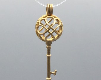 May include: A gold key-shaped pendant with a round, intricate, openwork design. The pendant is on a clear string.