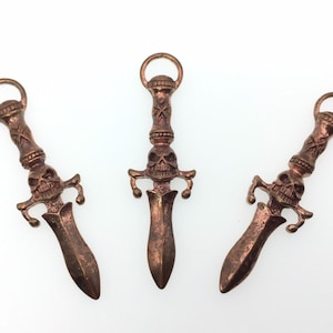 May include: Three copper-colored skull dagger charms with a detailed design. Each charm features a skull with a cross-hatched pattern on the handle and a pointed dagger blade.