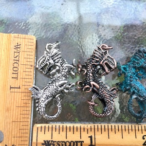 May include: Three silver dragon charms, each with a different finish. The charms are about 1 inch long and are shown next to a ruler for size reference.