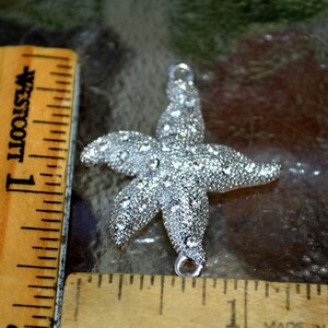 May include: Silver starfish charm with clear rhinestones. The charm is about 2 inches wide and has a loop at the top for attaching to a necklace or bracelet.