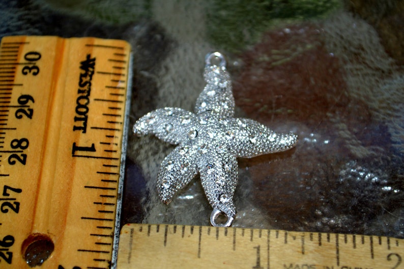 May include: Silver starfish charm with clear rhinestones. The charm is about 2 inches wide and has a loop at the top for attaching to a necklace or bracelet.