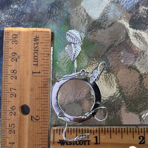 Puede incluir: Anillo de metal plateado con una abertura circular y dise&ntilde;os en forma de hoja. El anillo tiene un di&aacute;metro de aproximadamente 2,5 cm.