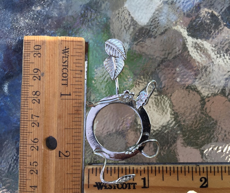 Puede incluir: Anillo de metal plateado con una abertura circular y dise&ntilde;os en forma de hoja. El anillo tiene un di&aacute;metro de aproximadamente 2,5 cm.