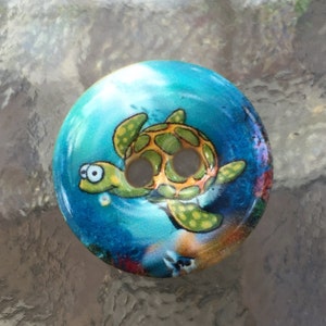 May include: A round wooden button with a colorful image of a sea turtle swimming in blue water with coral reefs. The turtle has green and yellow markings and big, round eyes.