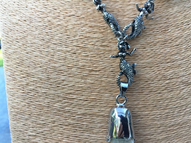 May include: A silver necklace with a bell pendant and multiple mermaid charms. The necklace is on a brown textured surface.