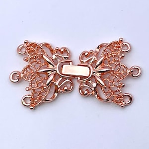 Puede incluir: Cierre de filigrana en oro rosa con diseño de mariposa. El cierre presenta intrincados patrones calados y pequeños bucles en cada extremo para la fijación. La barra central asegura las dos mitades. Un elemento decorativo para la fabricación de joyas.