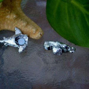 May include: Two silver charm pendants, one shaped like a shark and the other like a fish, both with a white stone in the center.
