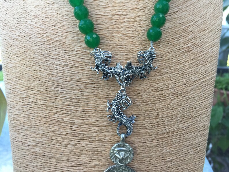 May include: A green jade bead necklace with a silver dragon pendant. The pendant features two dragons facing each other with a third dragon hanging below.