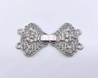 May include: Silver-toned metal clasp with a decorative filigree design and clear rhinestones. The clasp has two loops on each side for attaching to a garment.