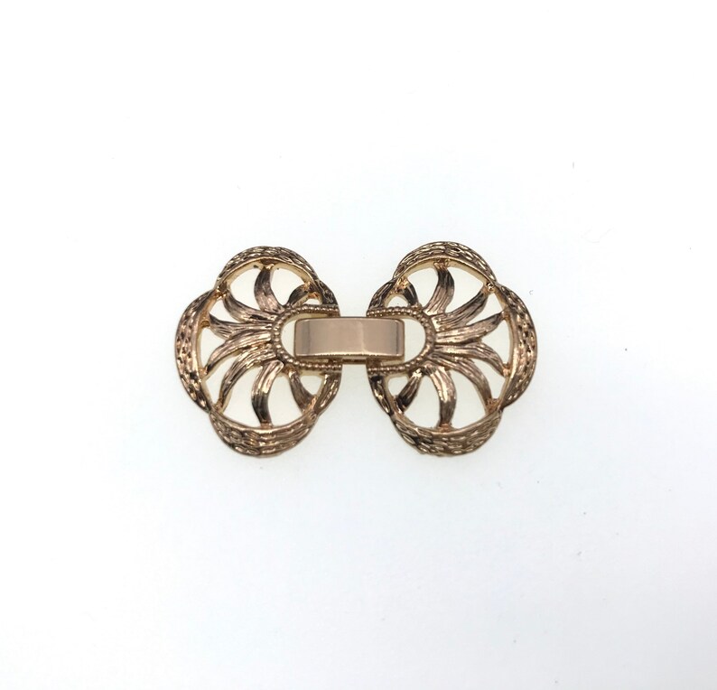 Puede incluir: Cierre de metal dorado con un dise&ntilde;o decorativo calado. El cierre tiene forma de dos c&iacute;rculos superpuestos con una barra central que los conecta.