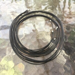 Puede incluir: Primer plano de un conjunto de collares de cordón negro con cierres plateados. Los collares están dispuestos en círculo sobre una superficie texturizada.