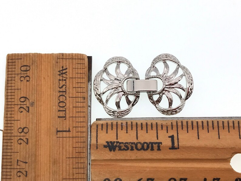 Puede incluir: Cierre de metal plateado con un dise&ntilde;o decorativo. El cierre mide aproximadamente 2,5 cm de ancho y se muestra junto a una regla.