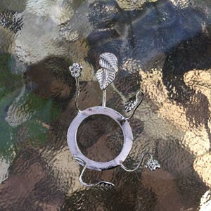 Puede incluir: Porta anillos de metal plateado con una base circular y tres brazos en forma de hoja. El porta anillos está diseñado para sostener un anillo u otro objeto pequeño.