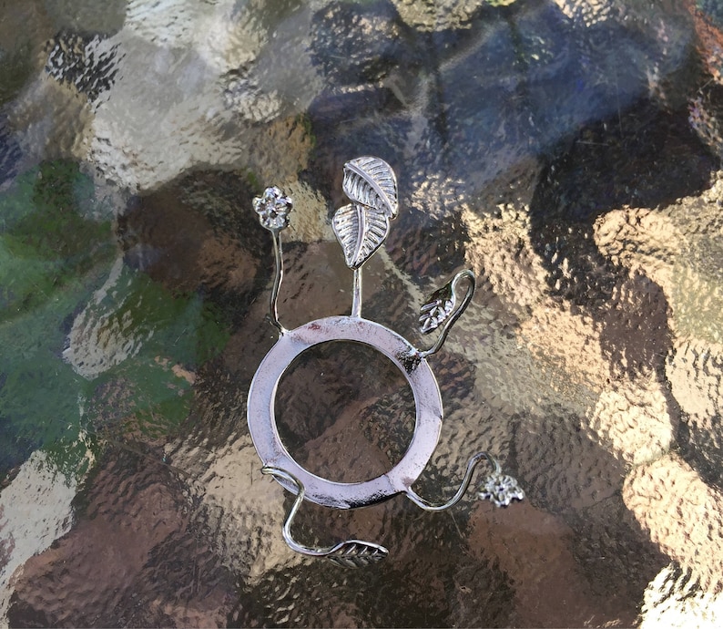 Puede incluir: Porta anillos de metal plateado con una base circular y tres brazos en forma de hoja. El porta anillos est&aacute; dise&ntilde;ado para sostener un anillo u otro objeto peque&ntilde;o.