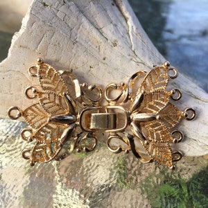 Puede incluir: Cierre de metal dorado con un diseño decorativo de mariposa. El cierre tiene dos bucles a cada lado y una barra central con un cierre de resorte.