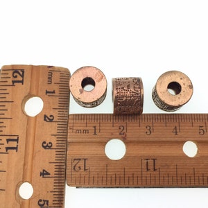 Puede incluir: Tres cuentas de metal de color cobre con dise&ntilde;os intrincados, medidas con una regla de madera. Las cuentas tienen aproximadamente 2,5 cm de di&aacute;metro.