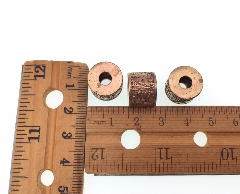 Puede incluir: Tres cuentas de metal de color cobre con dise&ntilde;os intrincados, medidas con una regla de madera. Las cuentas tienen aproximadamente 2,5 cm de di&aacute;metro.