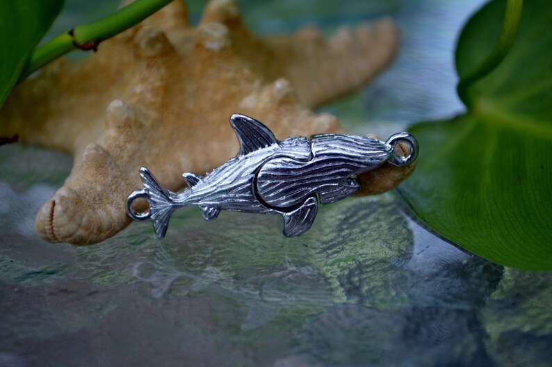 May include: A silver shark charm with a detailed, textured finish. The charm has a loop at the top for attaching to a necklace or bracelet.