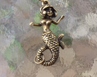 May include: A bronze-toned metal mermaid pendant featuring a detailed scale pattern on the tail, a curved body, and outstretched arms. The figure has a small loop at the top for attachment and a circular ring at the base of the tail fin.