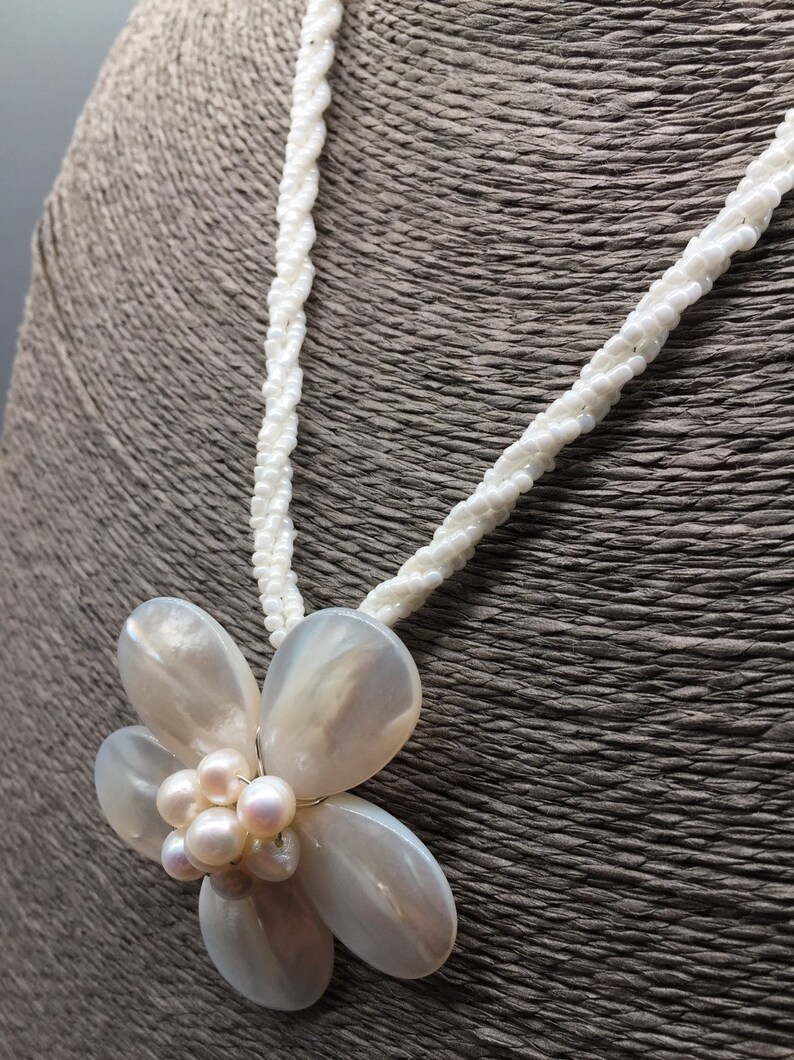 Pode incluir: Um colar de p&eacute;rolas brancas com um pingente de flor branca feito de concha. A flor tem um grupo de p&eacute;rolas no centro.
