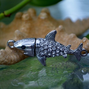 May include: A silver shark charm with rhinestones, perfect for adding a touch of whimsy to jewelry or crafts.