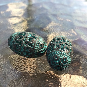May include: Two teal blue oval-shaped beads with intricate, carved designs. The beads have a distressed, aged look.