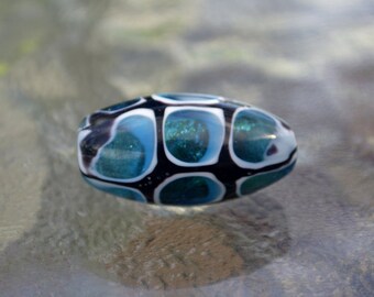 May include: A blue and black glass bead with white accents. The bead has a pattern of circles and swirls.