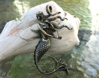 May include: A bronze mermaid brooch with a flowing tail and a crown on her head. She is holding a small object in her hand.