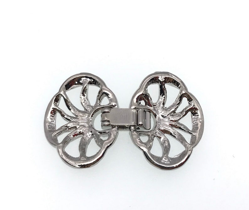 Puede incluir: Cierre de metal plateado con un dise&ntilde;o decorativo calado. El cierre tiene forma de dos c&iacute;rculos superpuestos con un patr&oacute;n floral detallado.