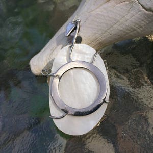 Puede incluir: Un colgante de n&aacute;car blanco con un marco de alambre plateado y una abertura circular en el centro. El colgante est&aacute; colgado de una cadena plateada.