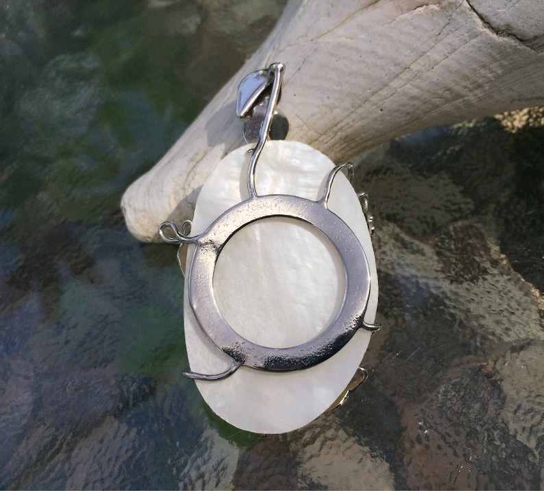Puede incluir: Un colgante de n&aacute;car blanco con un marco de alambre plateado y una abertura circular en el centro. El colgante est&aacute; colgado de una cadena plateada.
