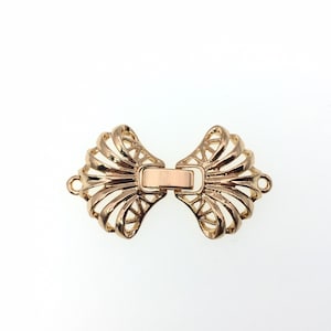 May include: Gold-toned metal clasp with a decorative, openwork design resembling a seashell. The clasp is designed to connect two pieces of jewelry or fabric.