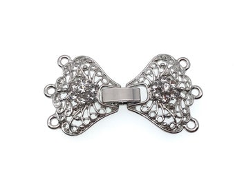 May include: Silver-toned metal clasp with a decorative filigree design and clear rhinestones. The clasp has two loops on each side for attaching to a necklace or bracelet.
