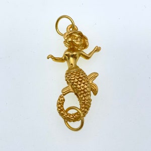 May include: A gold-colored mermaid charm with a detailed design. The mermaid has a crown and a long, flowing tail with scales. The charm has a loop at the top for attaching to a necklace or bracelet.