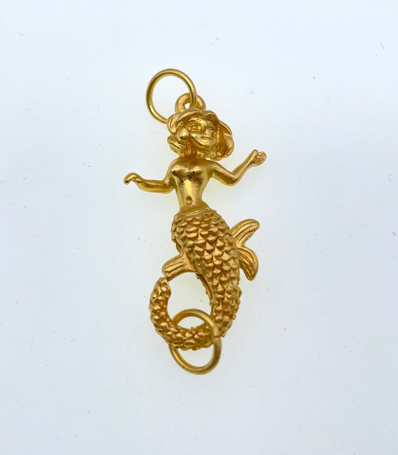 May include: A gold-colored mermaid charm with a detailed design. The mermaid has a crown and a long, flowing tail with scales. The charm has a loop at the top for attaching to a necklace or bracelet.
