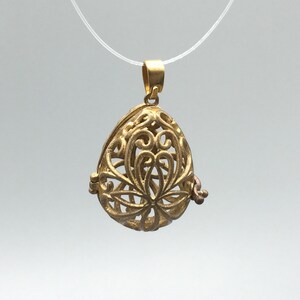 Może przedstawiać: Złoty wisiorek w kształcie jajka z filigranowymi zdobieniami, wiszący na przezroczystej sznurku.