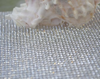 May include: A close-up of a white seashell resting on a sparkling silver background. The background is made of a grid of small, shiny, silver rhinestones.