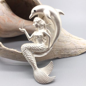 May include: A silver mermaid figurine with a dolphin tail, holding a small object in her hand. The figurine is sitting on a seashell.