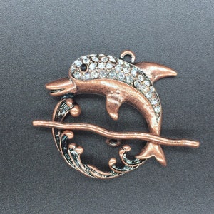 May include: A copper-toned dolphin charm with clear rhinestones, jumping over a wave design. The charm is perfect for jewelry making or crafting.