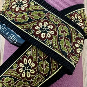 Puede incluir: Primer plano de una correa decorativa con un estampado floral. La correa presenta un fondo negro con un diseño repetido de flores y hojas burdeos y verde oliva. El texto "MAPLE & GRITS" es visible en una etiqueta negra.