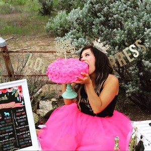 Puede incluir: Una mujer con una falda de tul rosa y una camiseta negra con un gr&aacute;fico de "Birthday Girl" de purpurina dorada est&aacute; comiendo un pastel glaseado rosa con un adorno de "Happy Birthday" de purpurina dorada. El pastel est&aacute; sobre un soporte para pasteles de color verde azulado. Hay una pancarta de purpurina dorada que dice "HOLLYWOOD PRESS" en el fondo.