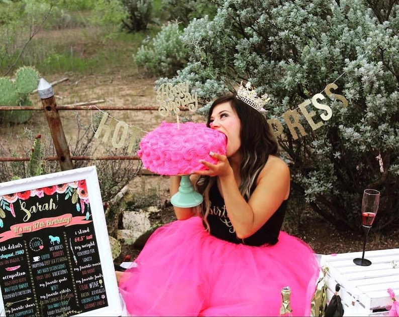 Puede incluir: Una mujer con una falda de tul rosa y una camiseta negra con un gr&aacute;fico de "Birthday Girl" de purpurina dorada est&aacute; comiendo un pastel glaseado rosa con un adorno de "Happy Birthday" de purpurina dorada. El pastel est&aacute; sobre un soporte para pasteles de color verde azulado. Hay una pancarta de purpurina dorada que dice "HOLLYWOOD PRESS" en el fondo.