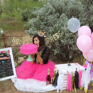 Puede incluir: Una mujer con una falda de tul rosa y una blusa de encaje negra sostiene un pastel rosa en forma de rosa frente a su cara. Lleva una corona dorada y una pancarta dorada con las palabras "Feliz cumplea&ntilde;os" detr&aacute;s de ella. Hay un cartel blanco con las palabras "Sarah 27th Birthday" en &eacute;l. Tambi&eacute;n hay globos rosas, blancos y dorados en el fondo.