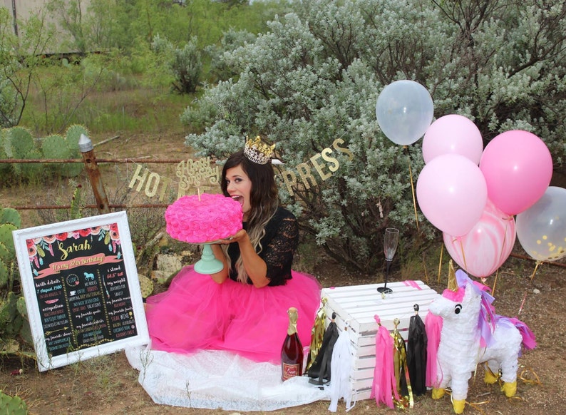 Puede incluir: Una mujer con una falda de tul rosa y una blusa de encaje negra sostiene un pastel rosa en forma de rosa frente a su cara. Lleva una corona dorada y una pancarta dorada con las palabras "Feliz cumplea&ntilde;os" detr&aacute;s de ella. Hay un cartel blanco con las palabras "Sarah 27th Birthday" en &eacute;l. Tambi&eacute;n hay globos rosas, blancos y dorados en el fondo.